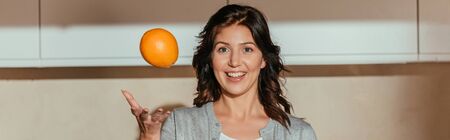 Panoramic crop of smiling woman throwing orange in kitchenの写真素材