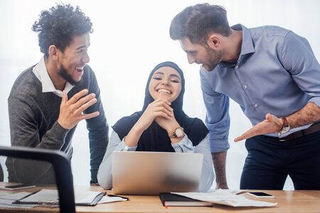 Selective focus of smiling muslim businesswoman near multicultural colleagues at table in office