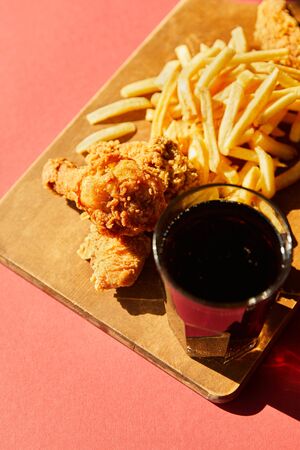 crispy deep fried chicken and french fries served on wooden cutting board with soda in sunlightの写真素材