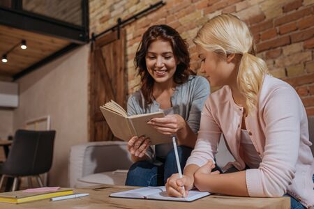 Smiling woman reading book near daughter writing on notebook at home