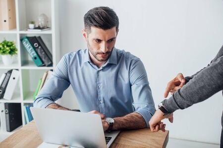 Businessman pointing at wristwatch to colleague near laptop on table in office