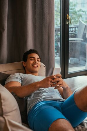 young happy mixed race man chilling and using smartphone on sofa during quarantineの写真素材