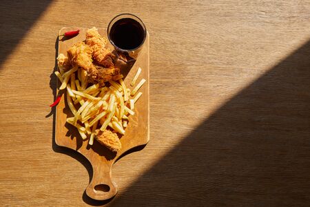 top view of spicy deep fried chicken, french fries on board with soda in glass on wooden table in sunlightの写真素材