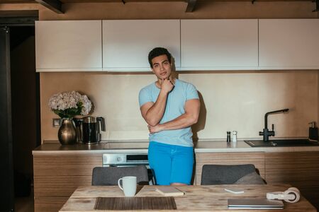 pensive mixed race man standing on kitchen with coffee cup, book, headphones and laptop during quarantineの写真素材