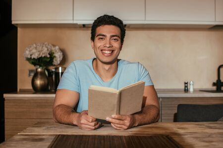 smiling mixed race man reading book on kitchen during quarantineの写真素材