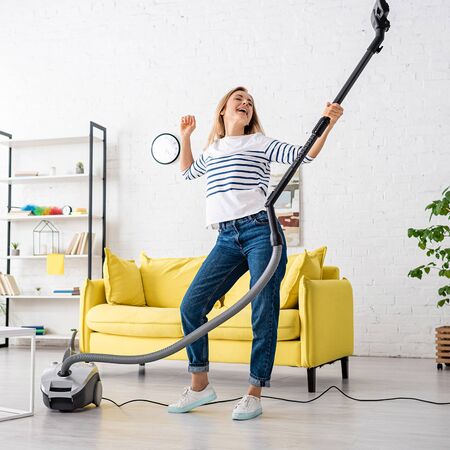 Blonde woman singing with closed eyes and vacuum cleaner near sofa in living roomの写真素材