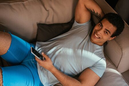 top view of happy mixed race man chilling and using smartphone on sofa during self isolationの写真素材