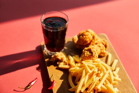 selective focus of spicy deep fried chicken and french fries served on wooden cutting board with soda in sunlightの写真素材