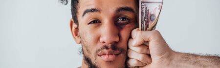 Male hand holding dollar banknotes near face of african american man with bruise isolated on grey, panoramic shotの写真素材