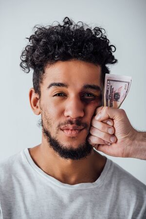 Male hand holding dollars near face of african american man with bruise isolated on grey