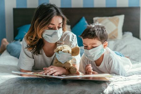 mother and toddler boy in medical masks reading book near soft toyの写真素材