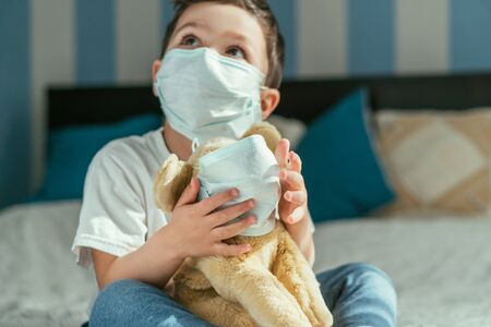 selective focus of toddler boy in medical mask holding soft toy and looking upの写真素材