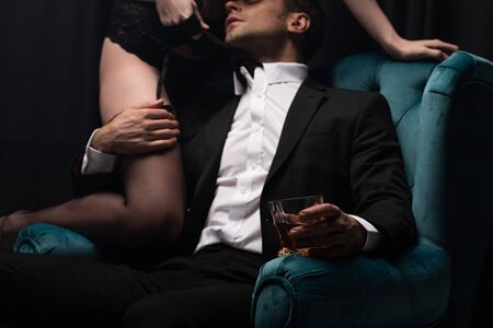cropped view of man in suit sitting in armchair, holding glass of whiskey and touching seductive...