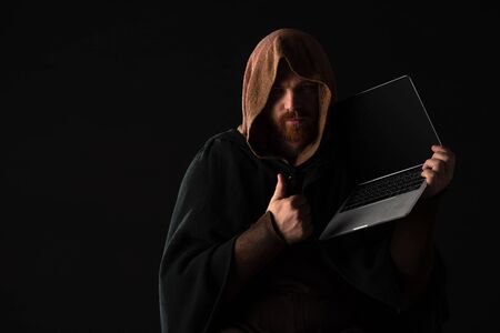medieval Scottish redhead man in mantel holding laptop and showing thumb up in dark isolated on...