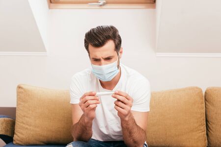 Man in medical mask holding thermometer on couch at home