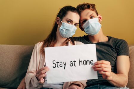 selective focus of man and woman in medical masks sitting on sofa and holding placard with stay at home letteringの写真素材