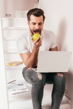 Handsome teleworker eating apple while working on laptop near documents on stairs