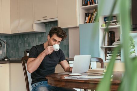 selective focus of handsome man drinking coffee and looking at camera while working at laptop in kitchenの写真素材