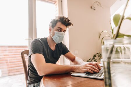 selective focus of young man in medical mask looking at camera while working on laptopの写真素材
