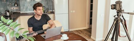 horizontal image of young vlogger playing guitar while looking at digital camera on tripod in kitchenの写真素材