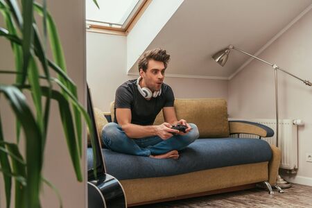 KYIV, UKRAINE - APRIL 13, 2019: selective focus of excited man in wireless headphones on neck playing video game while sitting on sofaの写真素材