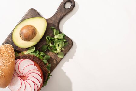 top view of delicious vegan burger with radish, avocado and greens on wooden cutting board on white...