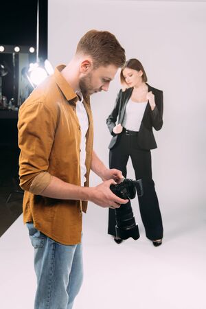 Selective focus of photographer looking at display of digital camera near elegant model and floodlight in photo studioの写真素材