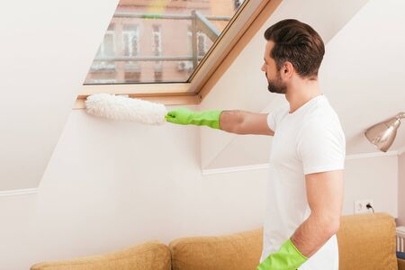 Side view of man in rubber gloves cleaning window with dust brush in living room
