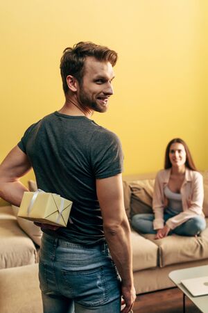 selective focus of man happy holding present behind back near girlfriend in living room