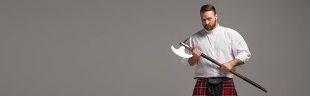 serious Scottish redhead man in red kilt with battle axe on grey background, panoramic shotの写真素材