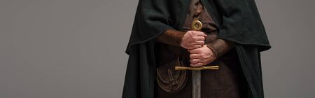 cropped view of medieval Scottish knight in mantel with sword in hands on grey background, panoramic...