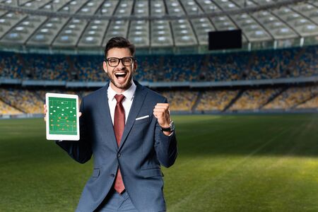 cheerful young businessman in suit and glasses holding digital tablet with soccer game on screen and showing yes gesture at stadiumの写真素材