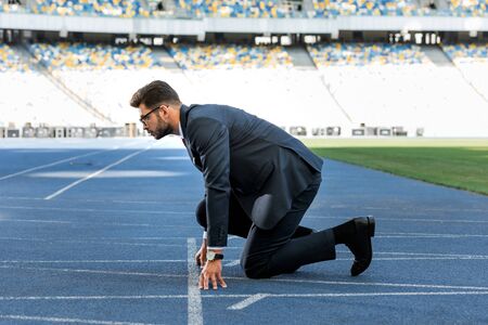 side view of young businessman in suit in start position on running track at stadium