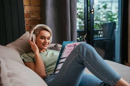smiling woman in wireless headphones reading magazine while resting on sofa at home