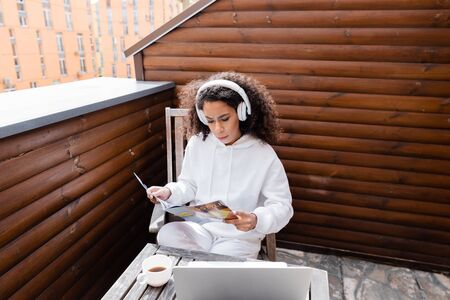 curly african american freelancer in wireless headphones reading magazine near laptop and cup of...