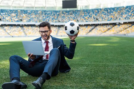 happy young businessman in suit with laptop and soccer ball sitting on football pitch at stadium, sports betting conceptの写真素材