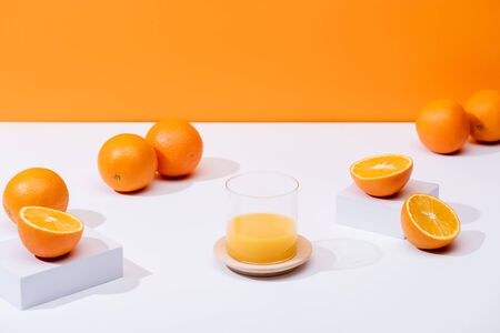fresh orange juice in glass near ripe oranges on white surface isolated on orange