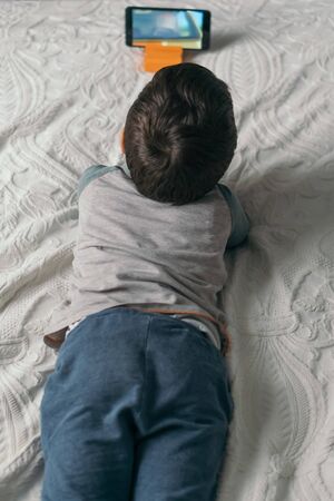 top view of little boy watching webinar on smartphone while lying on bedの写真素材