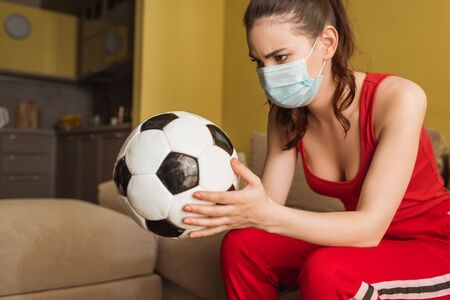 sportive woman in medical mask looking at football at homeの写真素材