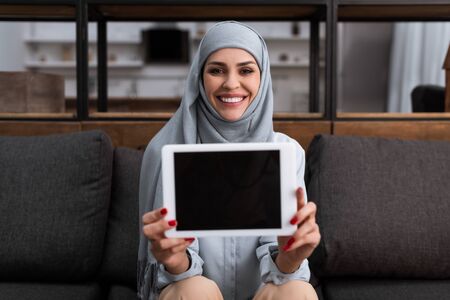 selective focus of smiling arabian woman in hijab holding digital tablet with blank screen in living roomの写真素材