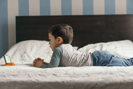 side view of cute boy lying on bed and watching online lesson on smartphoneの写真素材