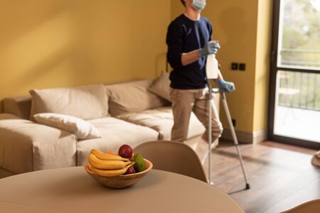Selective focus of disabled man in medical mask holding bottle of detergent and crutches in living roomの写真素材