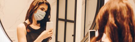 panoramic crop of woman in black dress and medical mask taking photo in bathroom