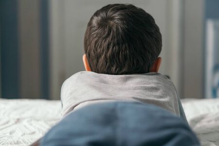 back view of little brunette boy lying on bed at homeの写真素材