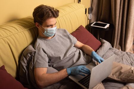 Man in medical mask and latex gloves working on laptop on bedの写真素材