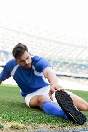 selective focus of professional soccer player in blue and white uniform stretching on football pitch at stadiumの写真素材