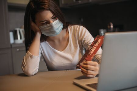 selective focus of sad freelancer in medical mask holding night party tickets near laptop at homeの写真素材
