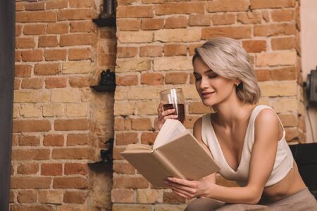 smiling woman holding glass of red wine while reading book at homeの写真素材