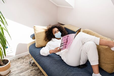 young african american woman in medical mask lying on sofa and reading magazine