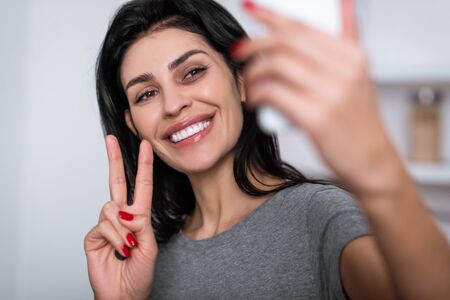 selective focus of smiling woman with bruise on face showing peace sign while taking selfie,...